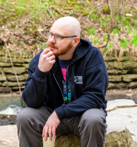 Photo of Chase Peterson. He is a bald bearded man wearing a CONvergence hoodie, sitting on a rock with his fist on his chin like he is thinking or listening to someone
