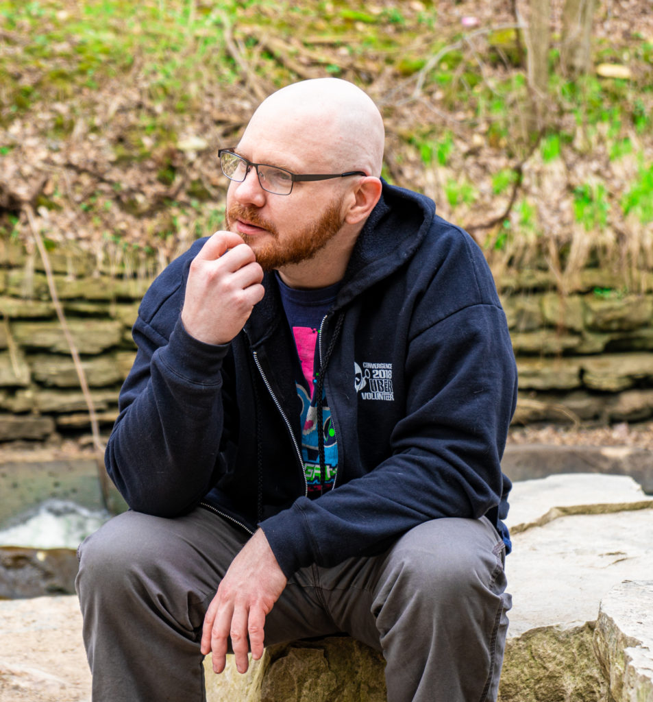 Photo of Chase Peterson. He is a bold bearded man wearing a hoodie, sitting with his fist on his chin like the thinker