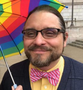 Photo of Eric Knight, a white man with a light beard and glasses. He is holding a rainbow umbrella and wearing a pink striped bow tie.