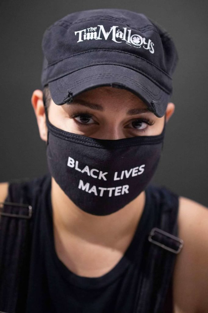 Photo of Lina DiGioia. She is wearing a black brimmed hat that says Tim Malloys on it, and a black face mask covering her nose and mouth that says Black Lives Matter on it. Only her eyes are visible. She is looking directly into the camera.