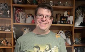 Photo of Tim Wick. He is a white man with brown hair and glasses. He is wearing a Star Wars shirt. He is smiling.