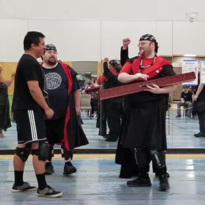 Photo of William Verburg-Cunningham at CONvergence 2019 in the Holodeck. He is talking with others, smiling, holding a long red box and is wearing a bandanna on his head.