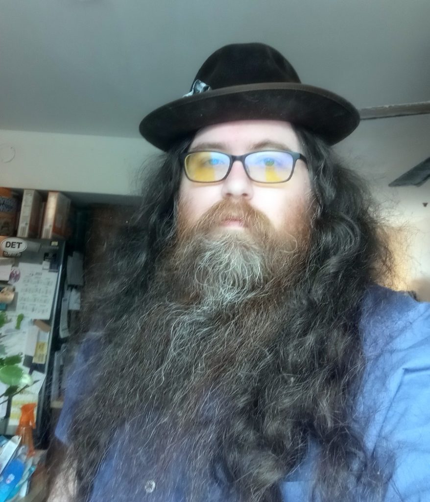 Headshot of Allen Tipper. They have glasses, are wearing a black hat, and have a large, brown beard with streaks of gray, framed by long brown hair over their shoulders.