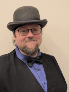 A photo of Will Verburg-Cunningham wearing a black coat, blue shirt, black bow tie, glasses and a gray hat.